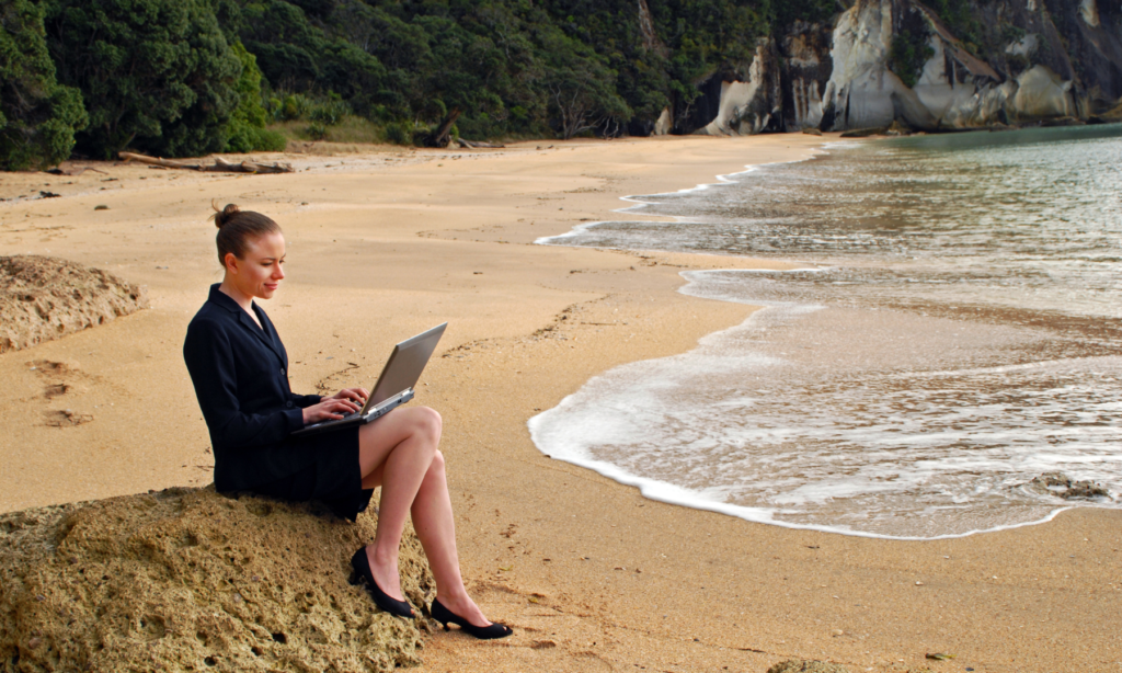 A woman sitting on a sand mound working on her laptop on the beach.