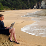 A woman sitting on a sand mound working on her laptop on the beach.