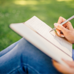 The side view of a woman writing in her notebook sitting in a patch of grass,