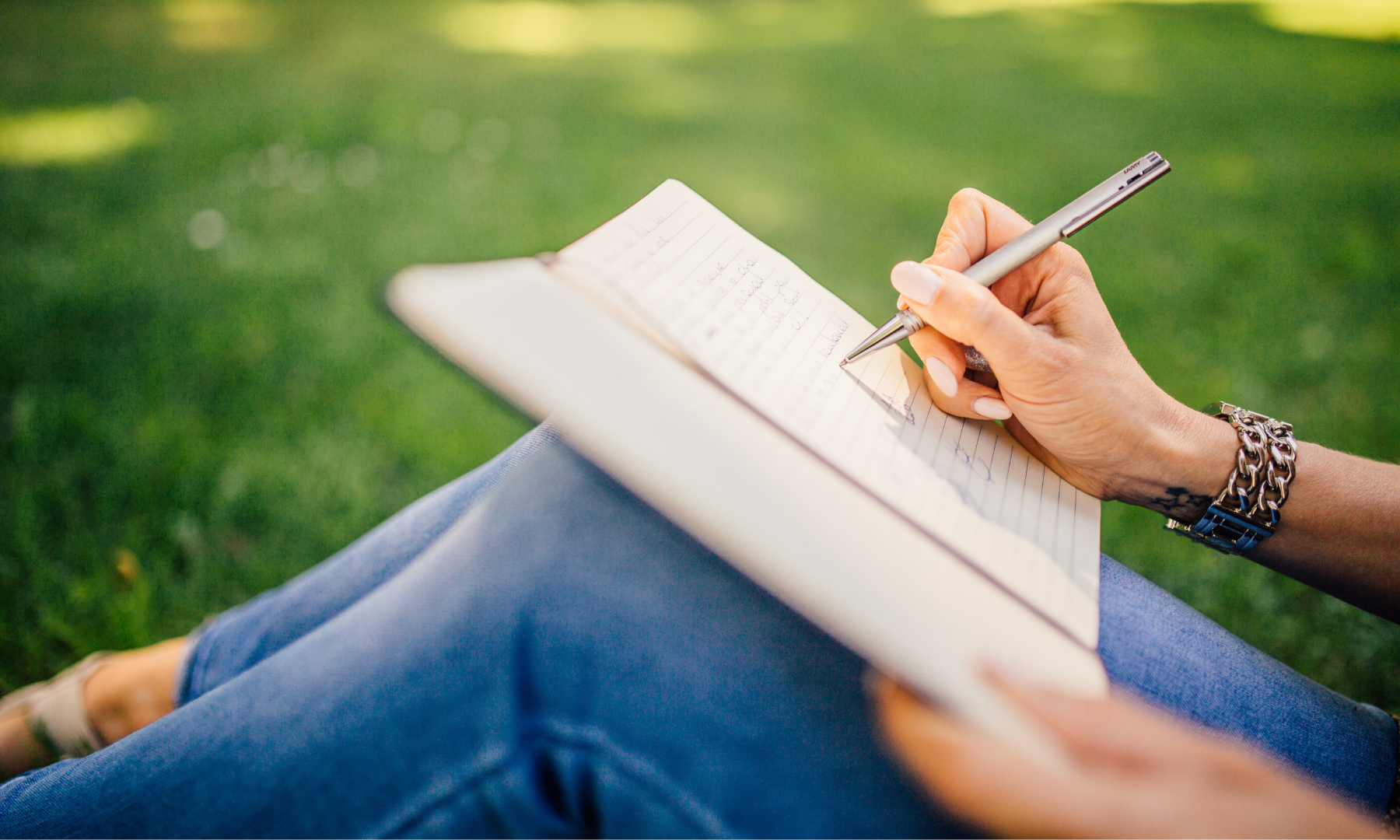 The side view of a woman writing in her notebook sitting in a patch of grass,